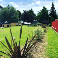 Un espace verdoyant bien entretenu, agrémenté de diverses plantes et arbres, avec un mur rouge et un bâtiment en arrière-plan.