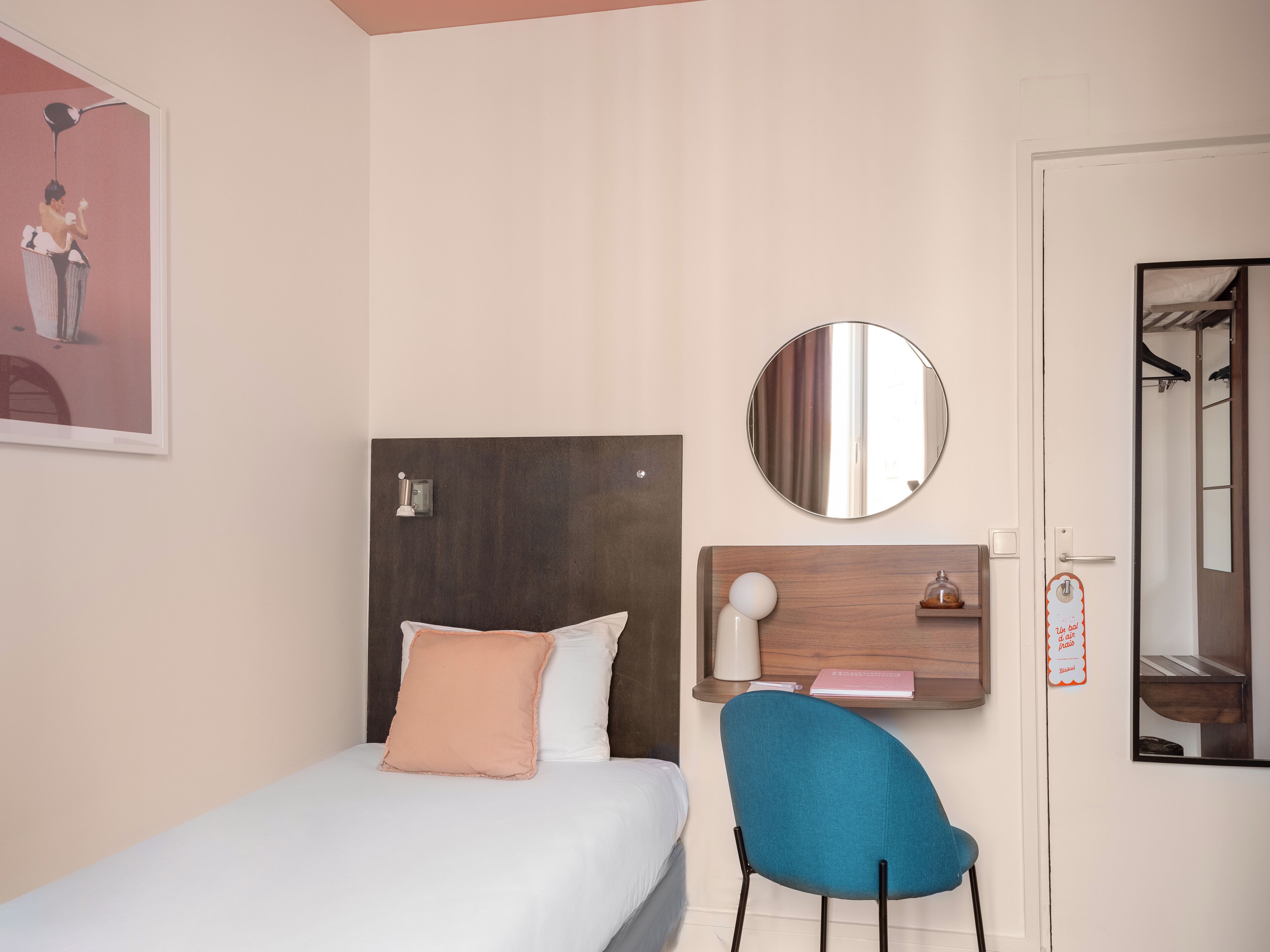 Un dormitorio con cama, una silla, un espejo redondo, un estante con un libro y un objeto pequeño, y una puerta con un letrero.