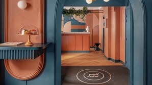 Un pasillo con pared azul, suelo de madera y un felpudo negro con un símbolo. Se observa una estantería con una campana de cristal y un libro.