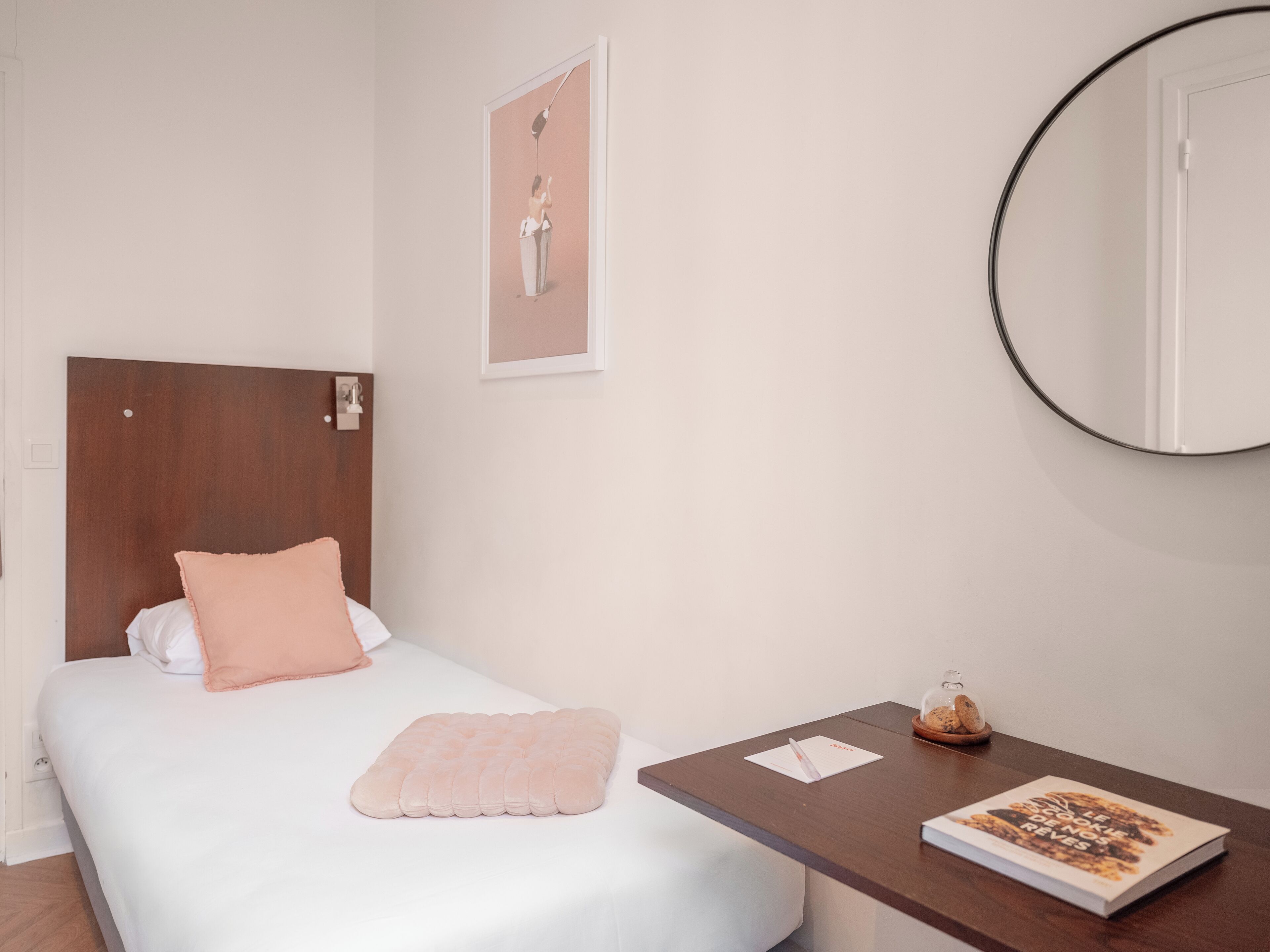 Un dormitorio ordenado con una cama, una mesita de noche con un libro y un recipiente de vidrio, un cuadro enmarcado en la pared y un espejo redondo.