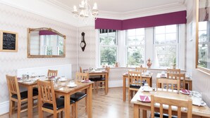 A dining area with wooden tables and chairs, a chandelier, and large windows with a view of the city. - The Ravenswood B&B (Torquay)
