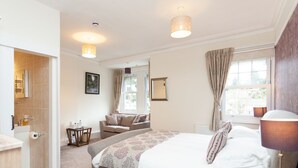 A bedroom with a large bed, a bedside table, a nightstand, a window with curtains, a bathroom with a mirror and towel rack, and a small table with a lamp. - The Ravenswood B&B (Torquay)