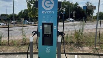 Una estación de carga EV Zen con una capacidad de 240 kW, ubicada en un estacionamiento con cielo despejado y algunos árboles al fondo.