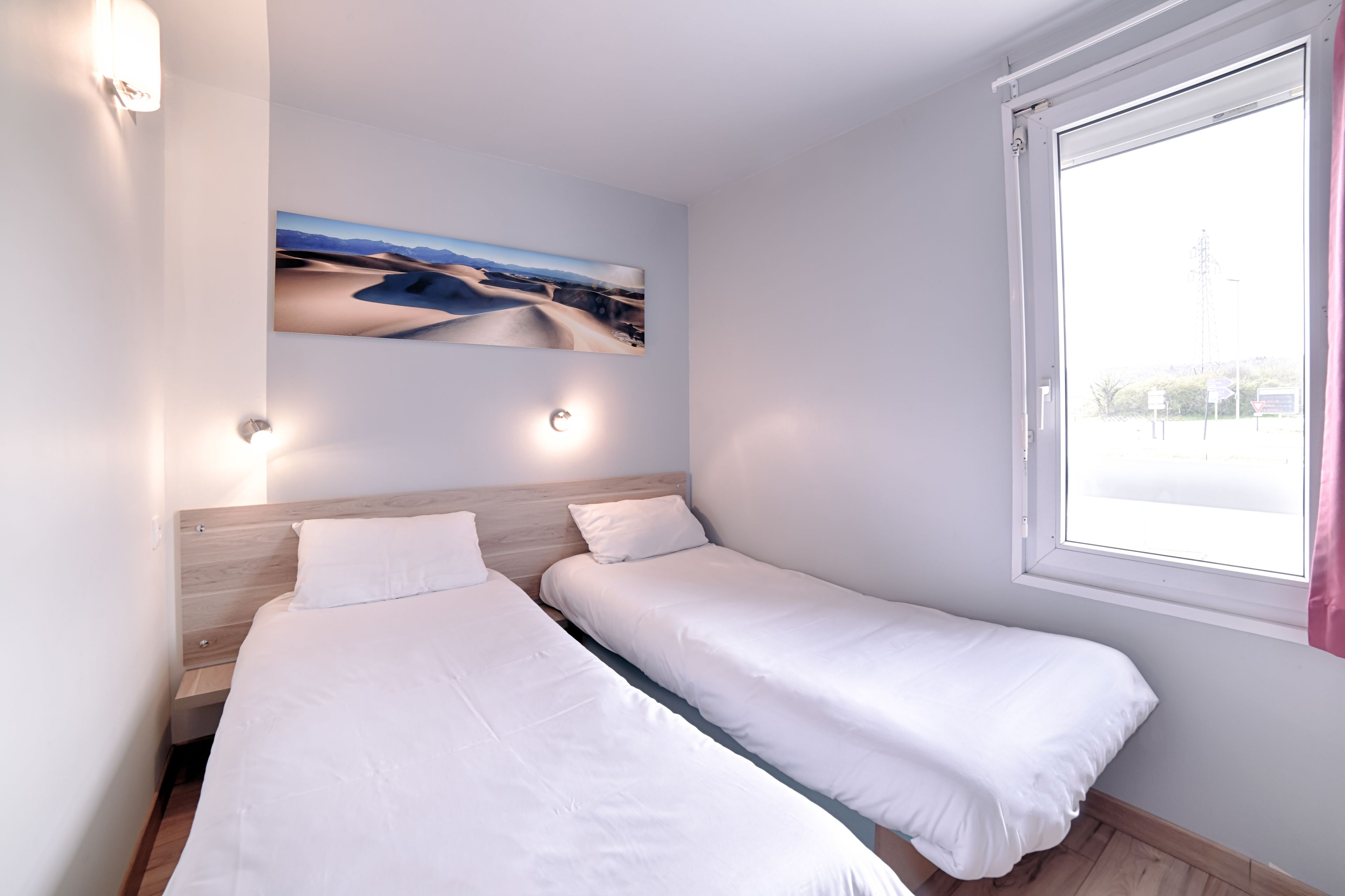 A room with two single beds, a wooden headboard, a wall-mounted artwork, and a window with a view of the city.