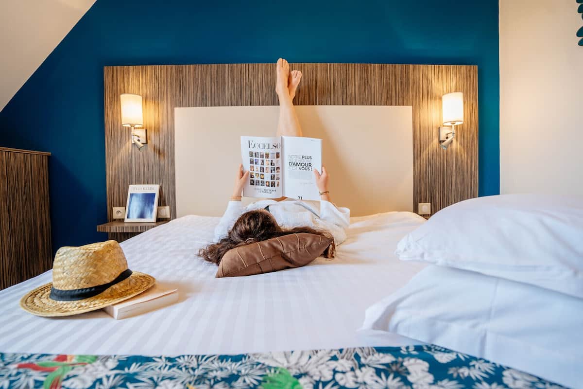 A person lying on a bed with a book, a hat, and a framed picture on a bedside table.