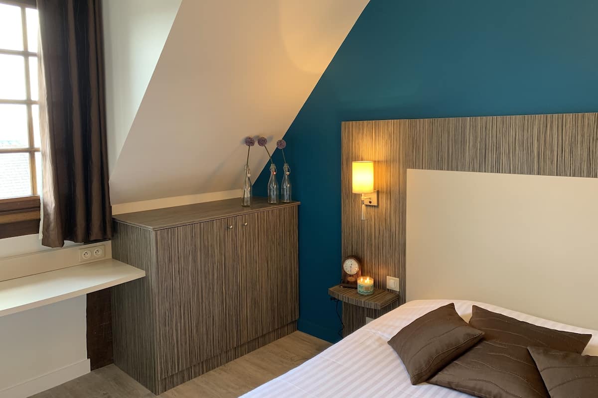 A bedroom with a sloped ceiling, a wooden headboard, a bedside table with a lamp, and a bed with multiple pillows.
