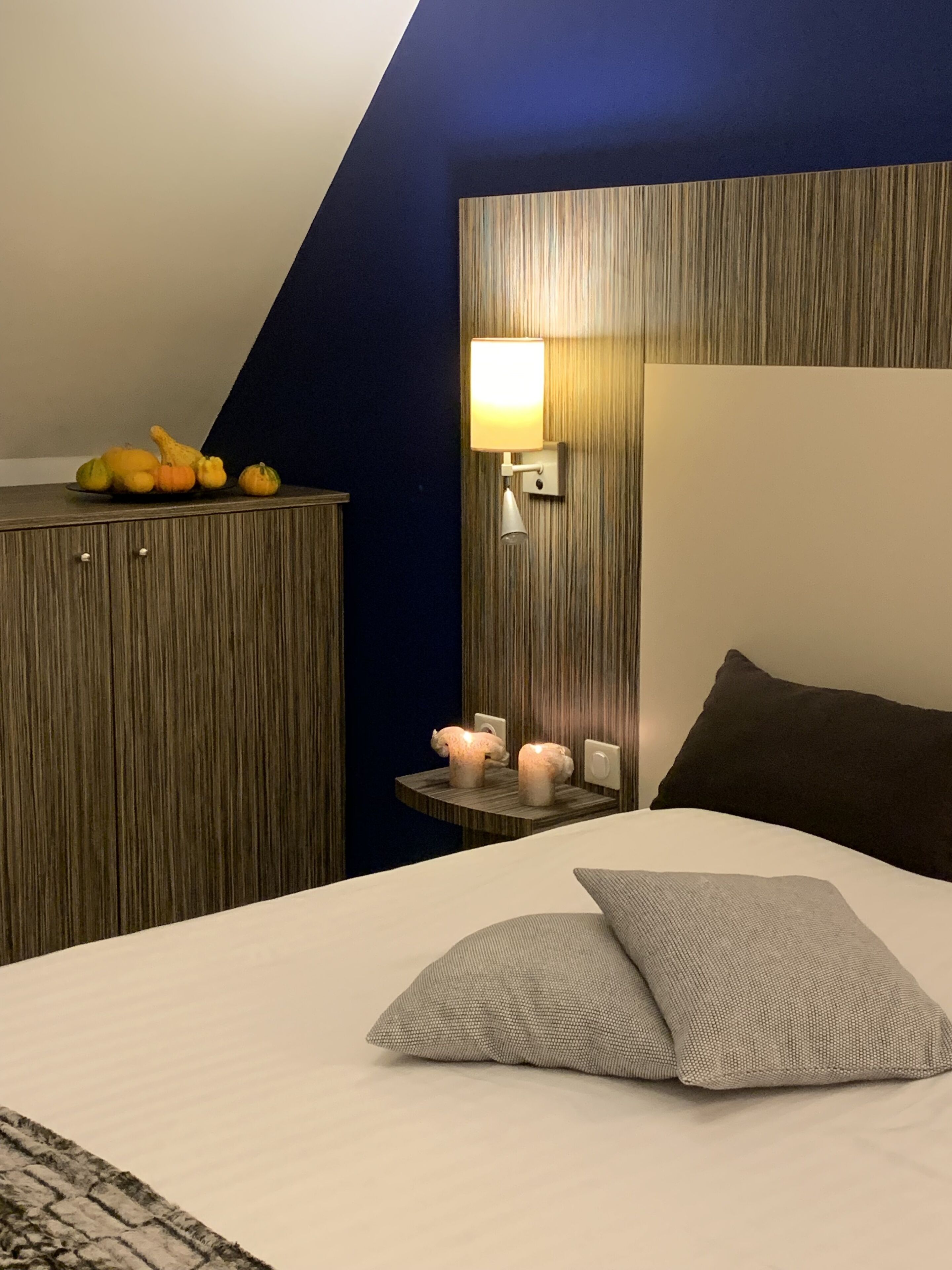 A hotel room with a bed, a nightstand with a lamp, a wooden headboard, and a wall-mounted shelf with decorative items.