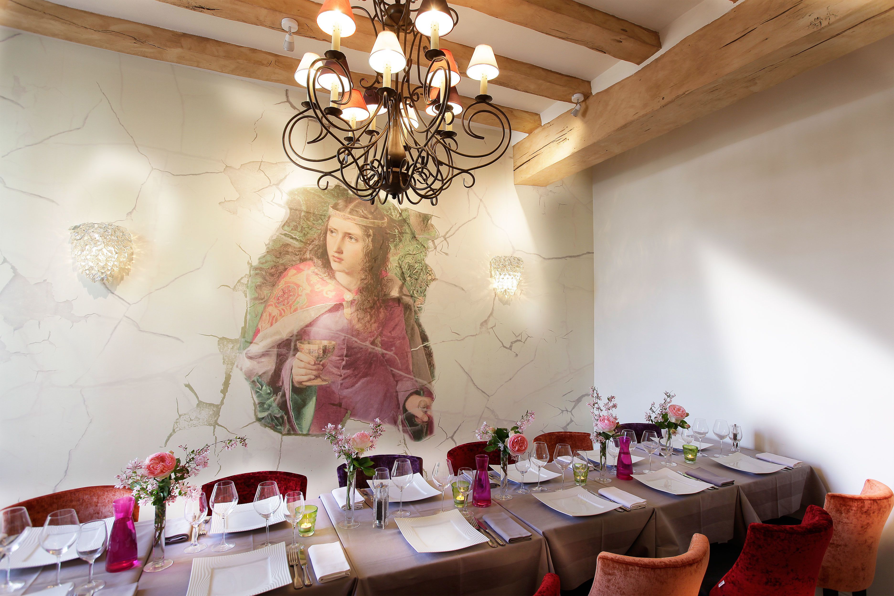 Un comedor con una larga mesa dispuesta para una comida, una araña y un mural en la pared.