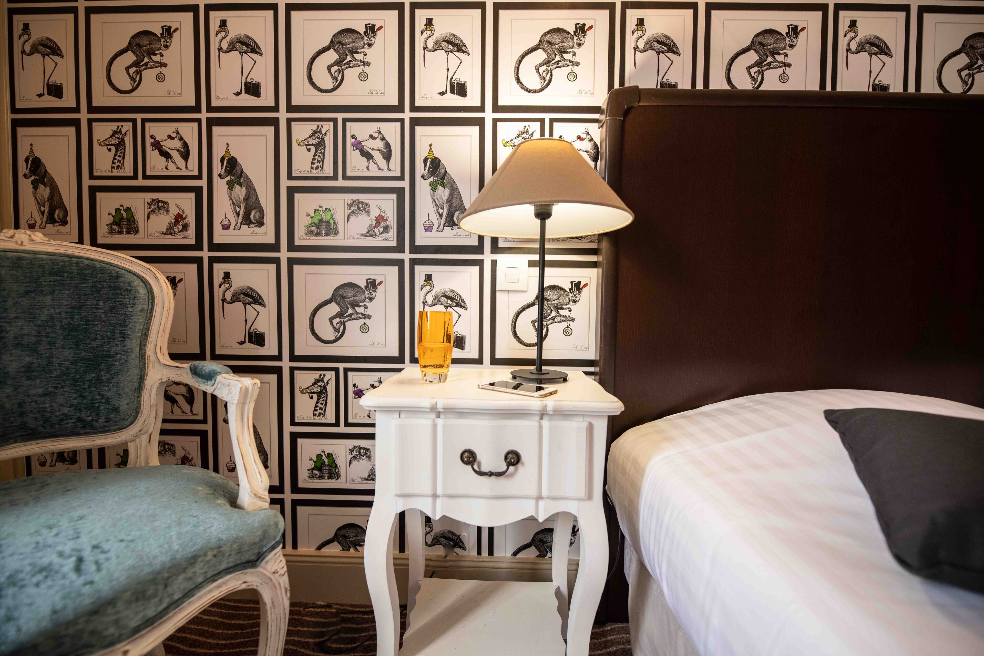 Une chambre d’hôtel comprenant un lit, une chaise, une table de chevet avec une lampe, et un mur décoré d’illustrations d’animaux encadrées.