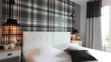 A hotel room with a plaid-patterned wall, a tufted headboard, a bedside table with a lamp, a bed with white bedding and a black pillow, and a window with curtains.