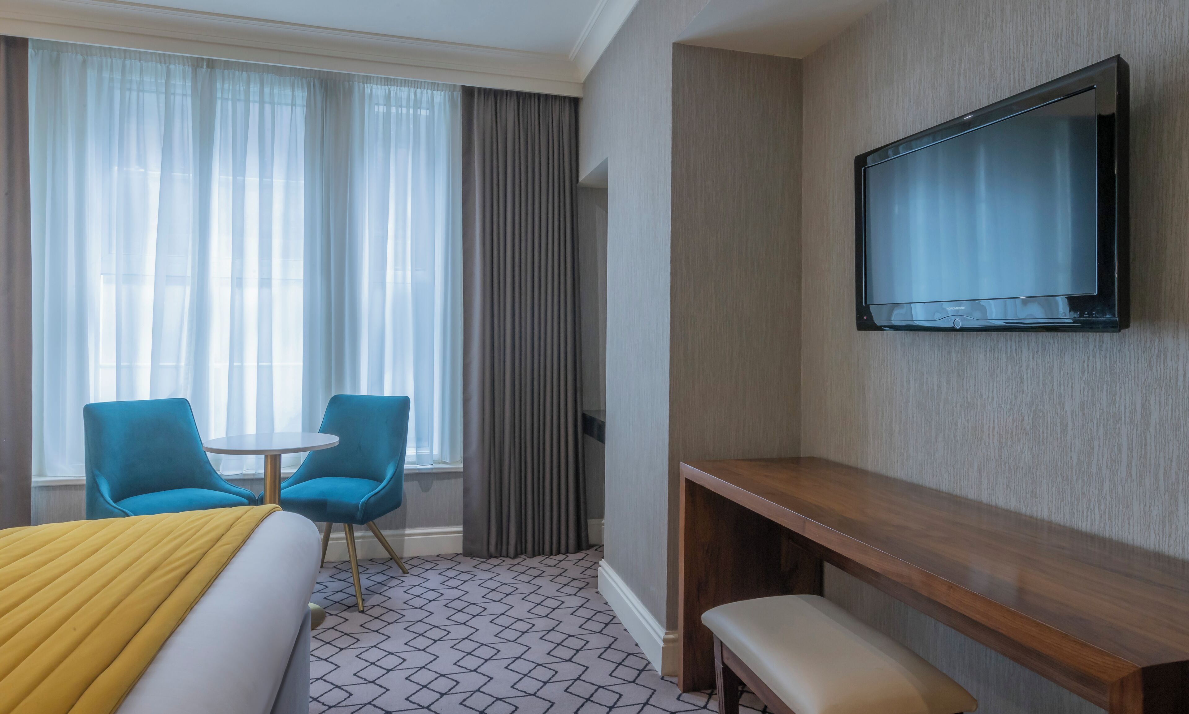 A hotel room with a bed, two blue chairs, a small table, a TV mounted on the wall, and a patterned floor.