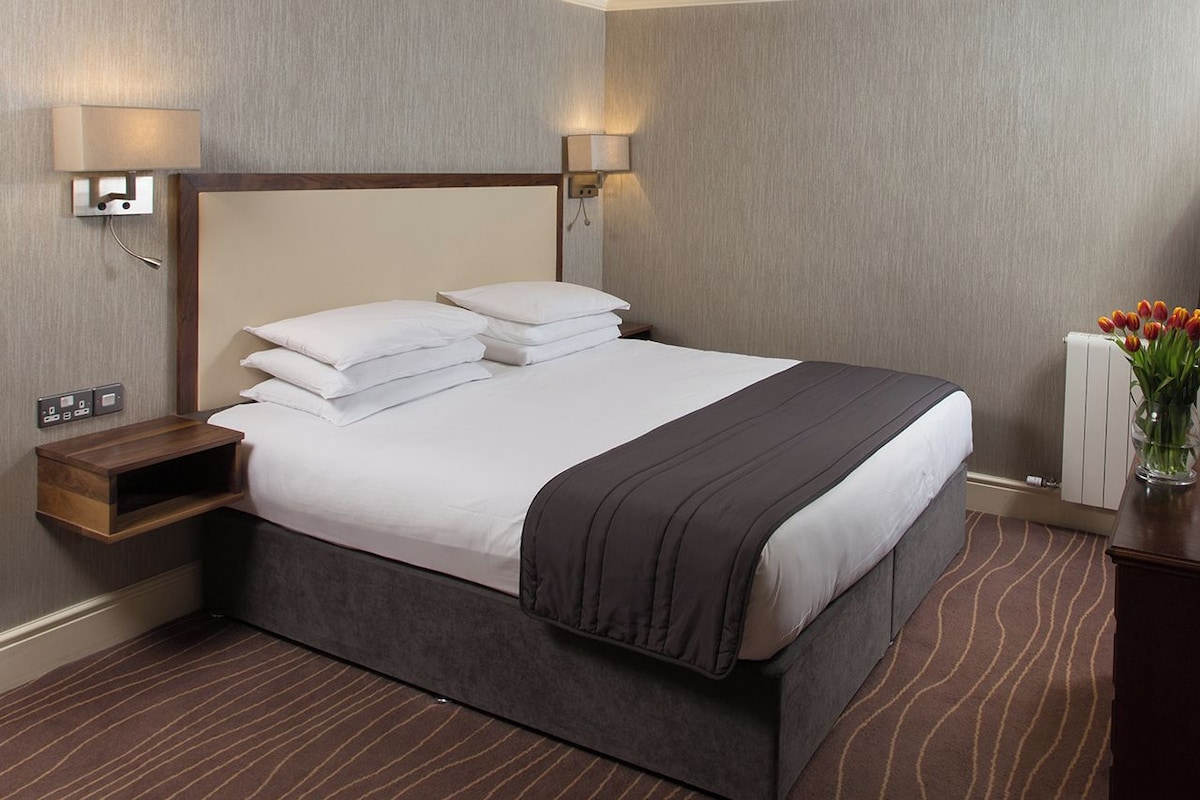 Habitación de hotel con una cama grande, dos lámparas de noche, una mesita de noche, un jarrón con flores y un radiador.