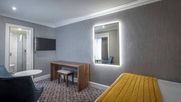 A modern hotel room with a wooden desk, a chair, a small round table, a bed with a yellow bedspread, a large mirror, and a flat-screen TV mounted on the wall.