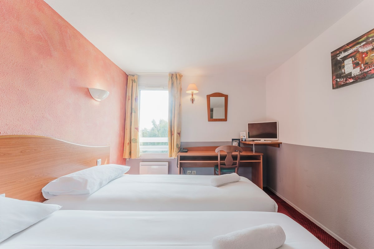 A hotel room with two beds, a desk, a chair, a television, and a window with curtains.