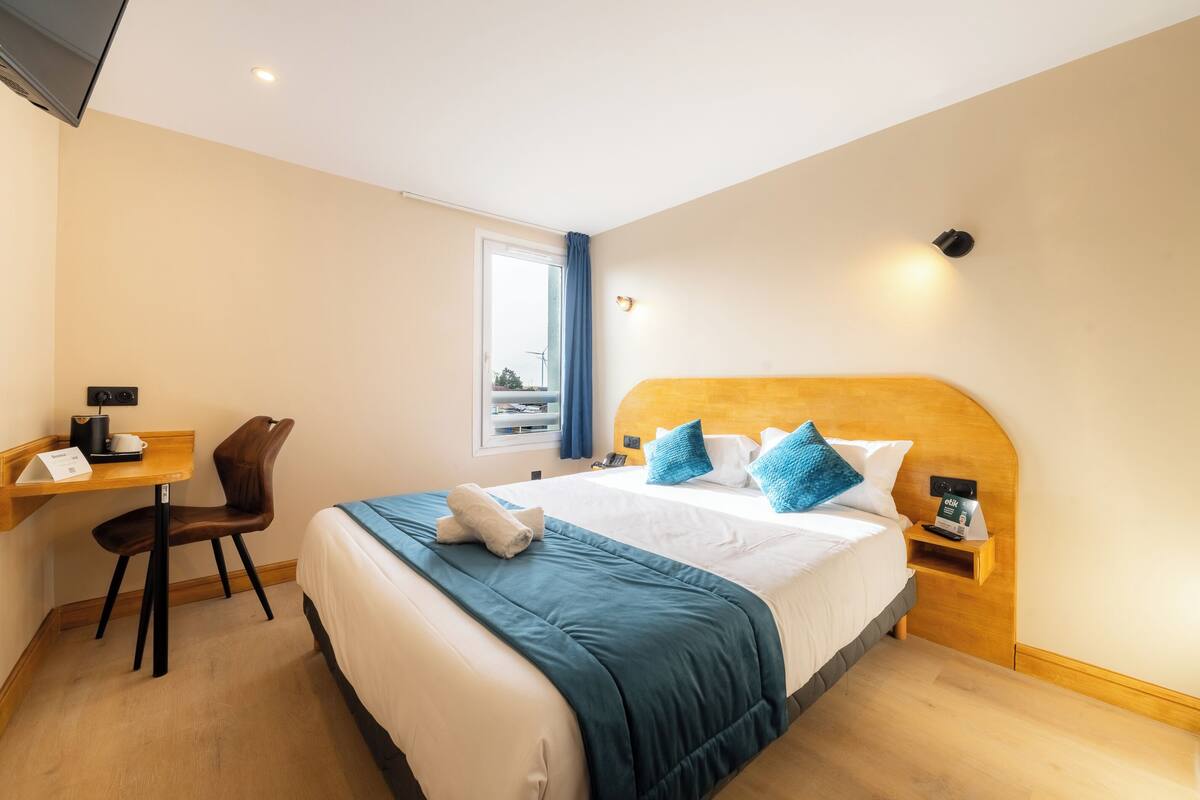 A hotel room with a bed, a desk, a chair, a nightstand, and a window.
