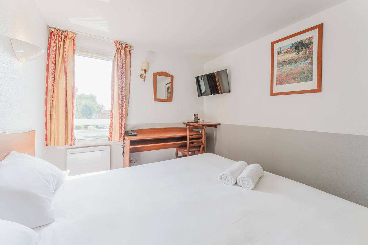 A hotel room with a bed, a desk, a chair, a TV, and a window with curtains.