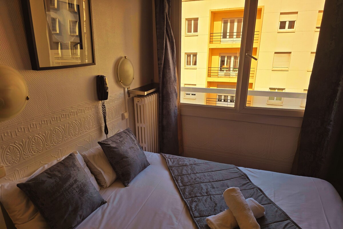 Une chambre d’hôtel équipée d’un lit, d’oreillers, d’un téléphone sur la table de chevet, d’un radiateur et d’une fenêtre donnant sur un immeuble.
