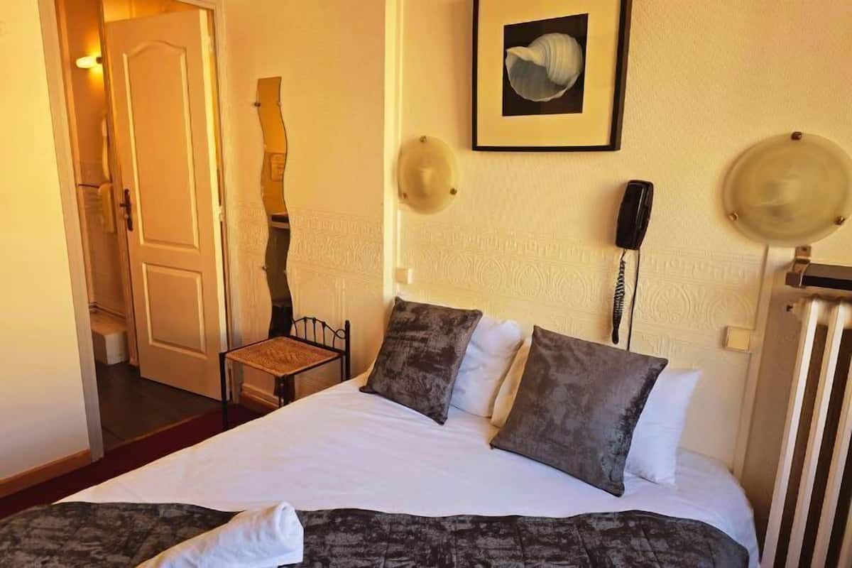 Ein Hotelzimmer mit einem Bett, einem Sessel, einer Tür, einem gerahmten Bild an der Wand, einem Telefon und einer Lampe.