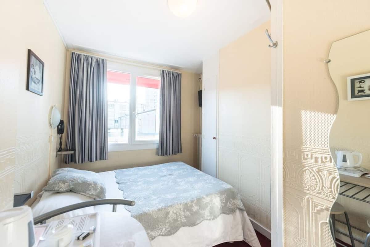 Ein Hotelzimmer mit Bett, Nachttisch, Stuhl, Fenster mit Vorhängen und einer Tür mit dekorativer Verkleidung.