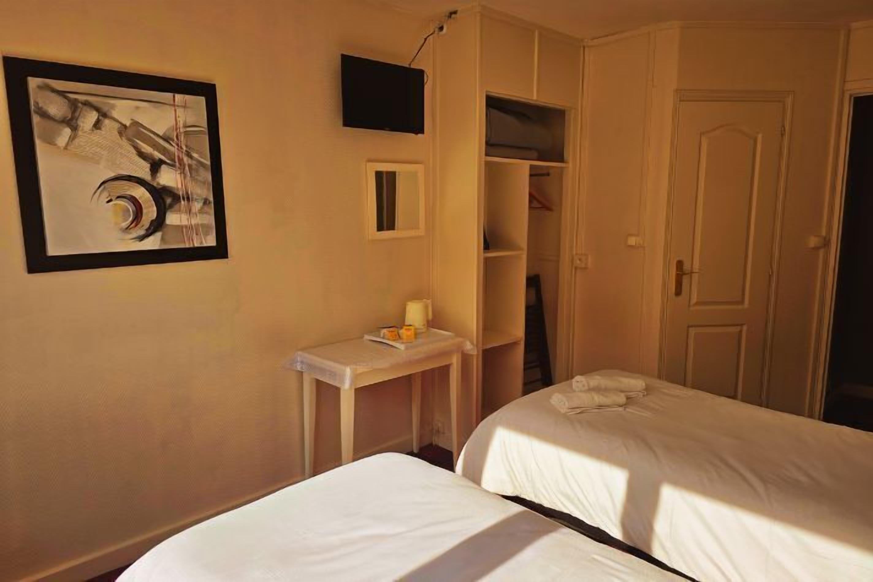 A hotel room with two beds, a small table with a lamp, a painting on the wall, and a wardrobe.