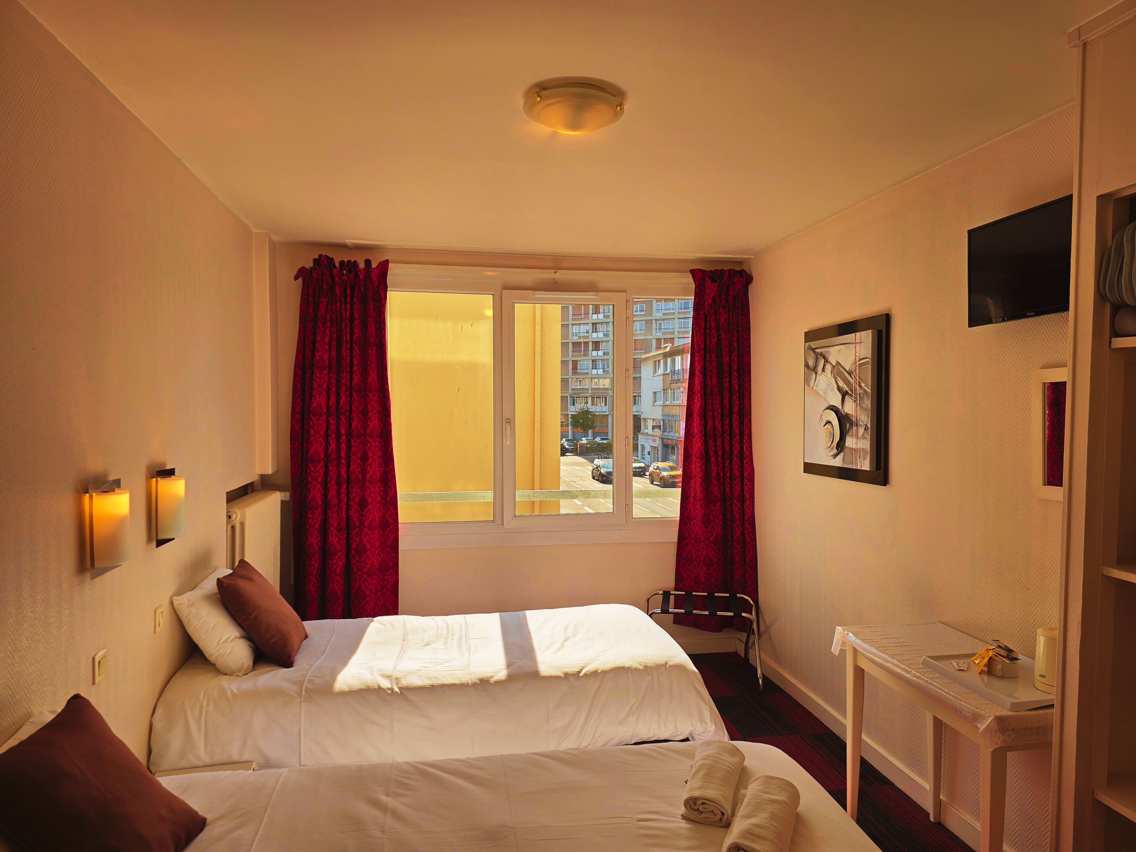 A hotel room with a bed, a window with curtains, a wall-mounted TV, and a small table with a cup and tissues.