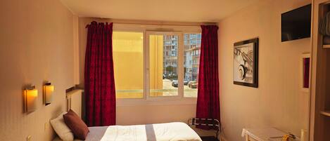 A hotel room with a bed, a window with curtains, a wall-mounted TV, and a small table with a cup and tissues.