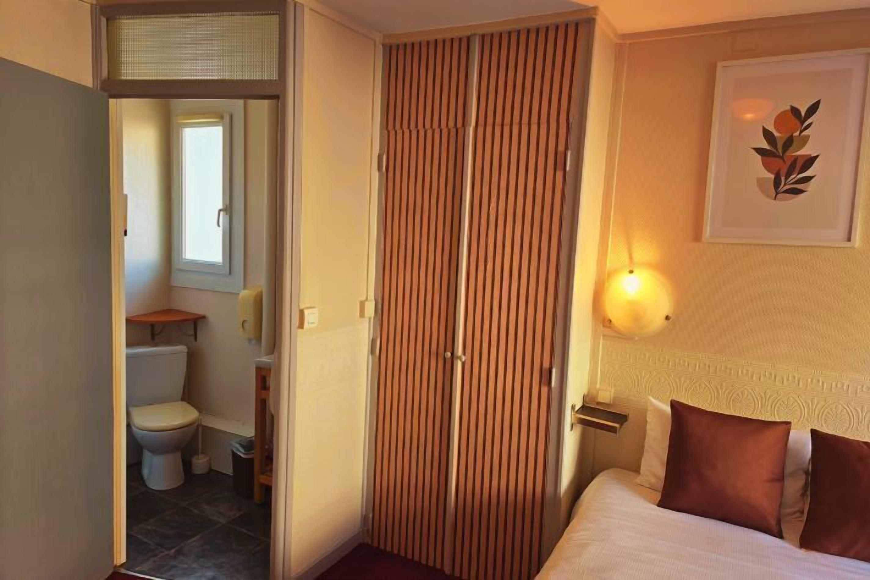Ett hotellrum med en säng, en garderob i trä, ett badrum med toalett och handfat samt ett inramat konstverk på väggen.