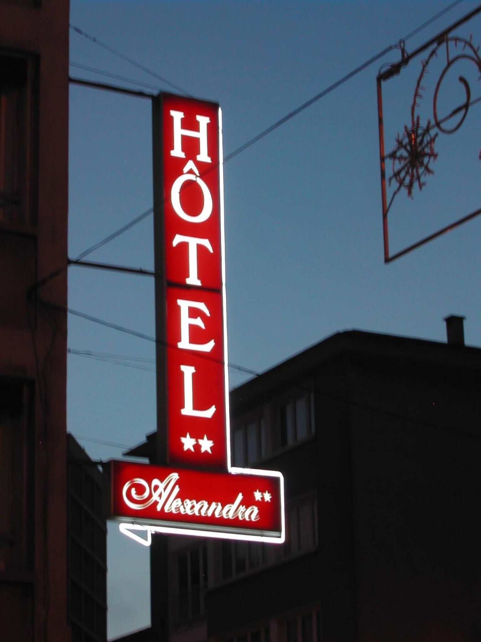 Eine vertikal leuchtende Neonreklame für das Hotel ‚Hôtel Alexandra‘ mit einem Sternsymbol unter dem Namen.