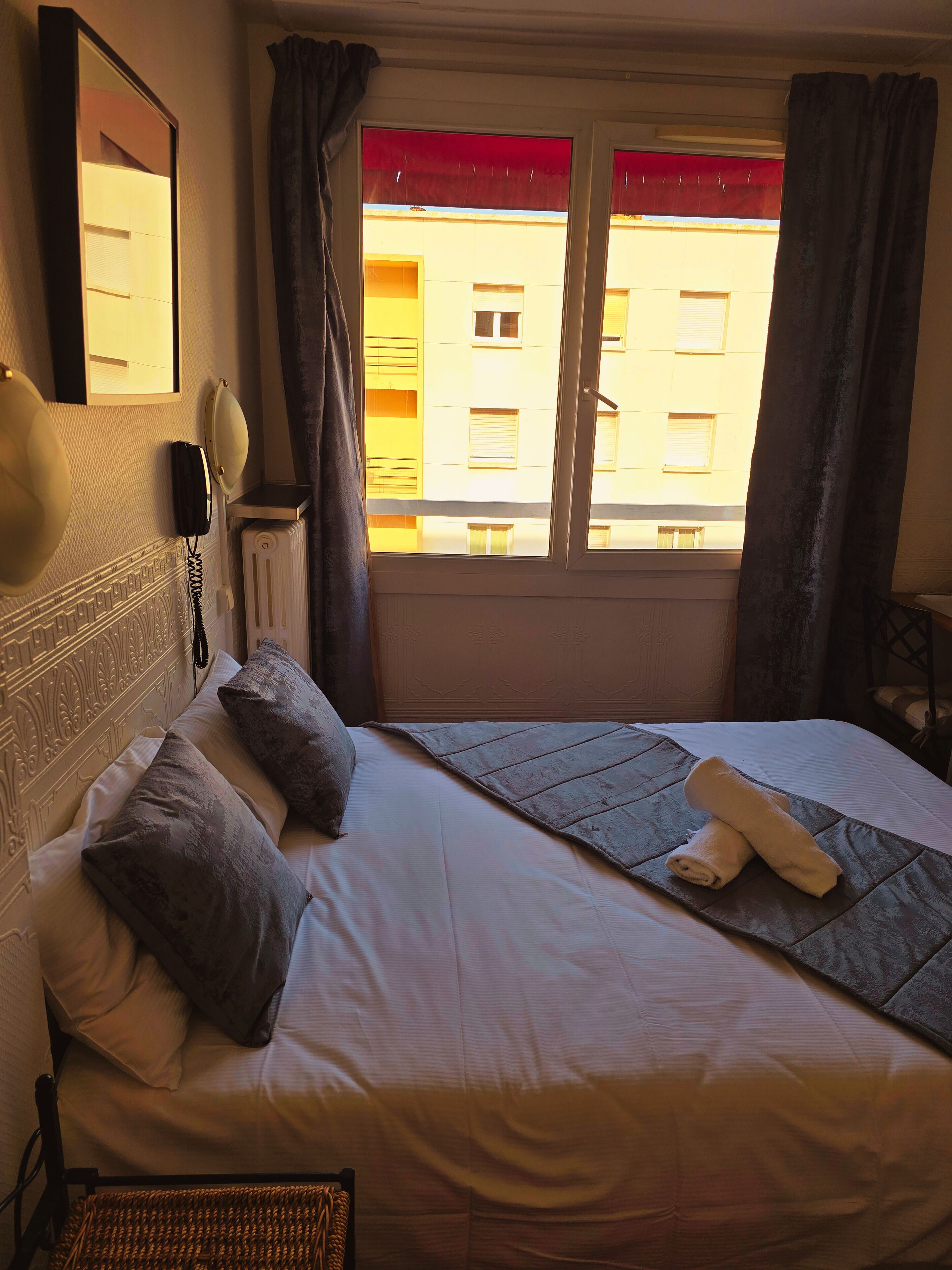 A hotel room with a bed, a telephone, a radiator, and a window with curtains.