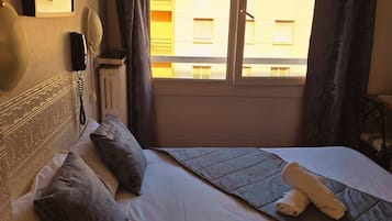 A hotel room with a bed, a telephone, a radiator, and a window with curtains.