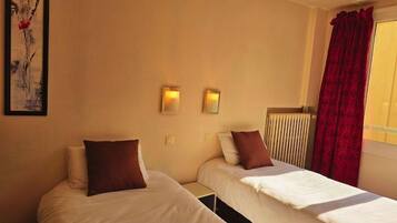 A hotel room with two single beds, a radiator, a red curtain, and a painting on the wall.