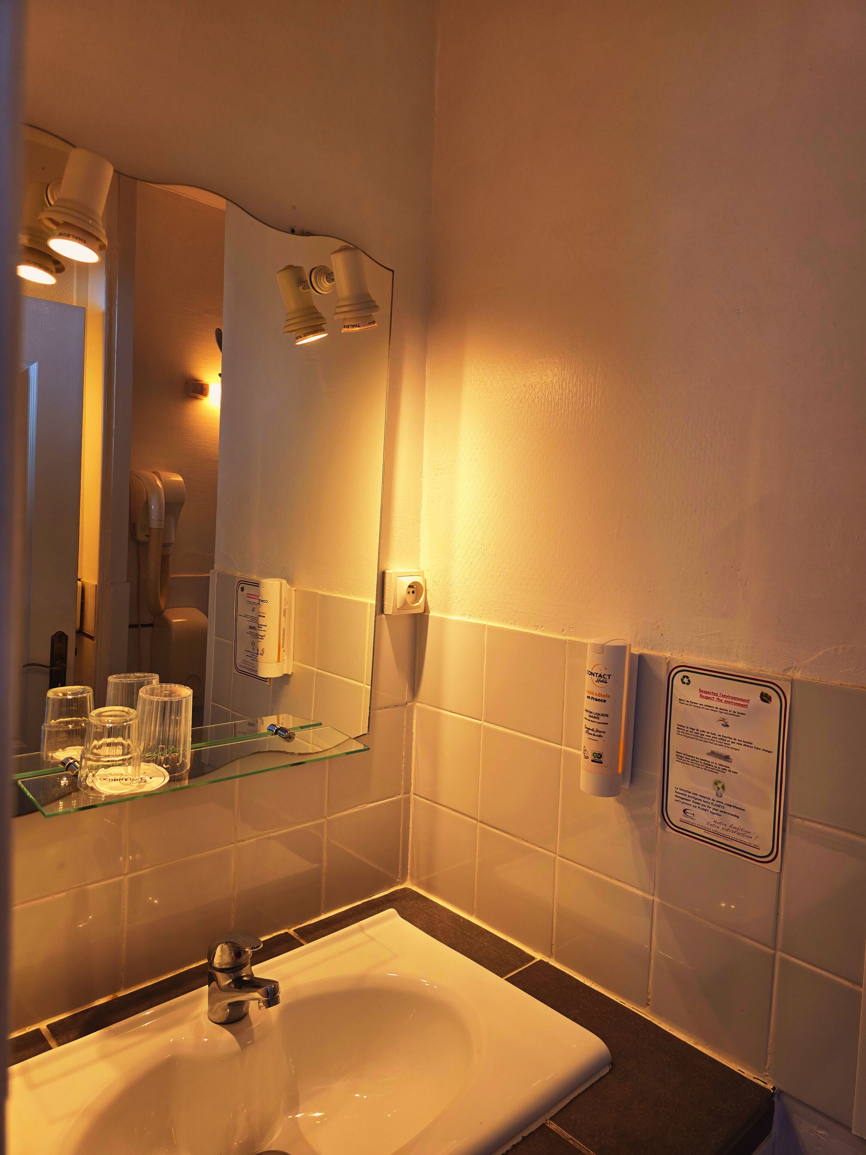 A bathroom with a sink, mirror, and glass shelves. There is a sign on the wall and a towel dispenser.