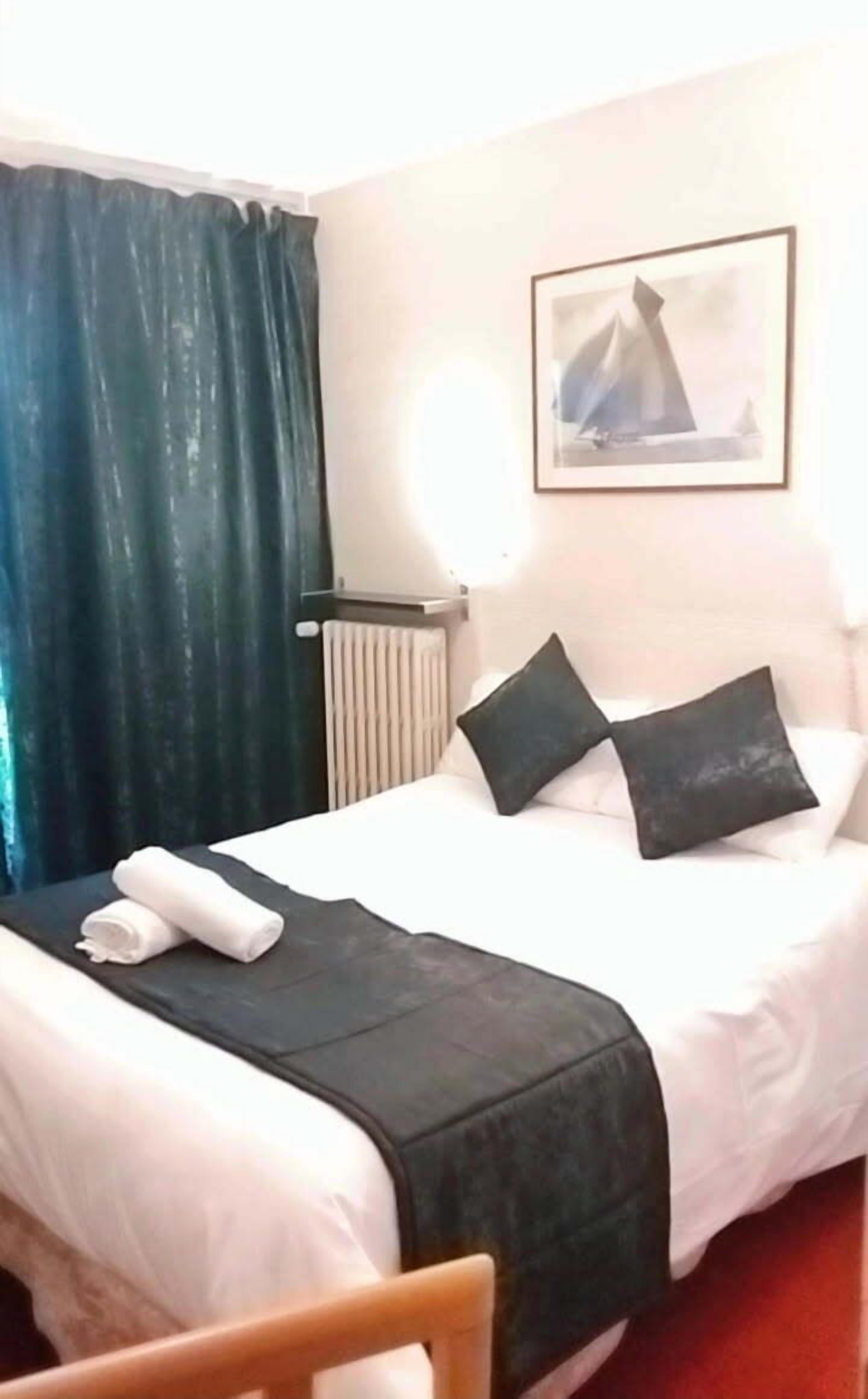 A hotel room with a bed, a radiator, a window with curtains, and a painting on the wall.
