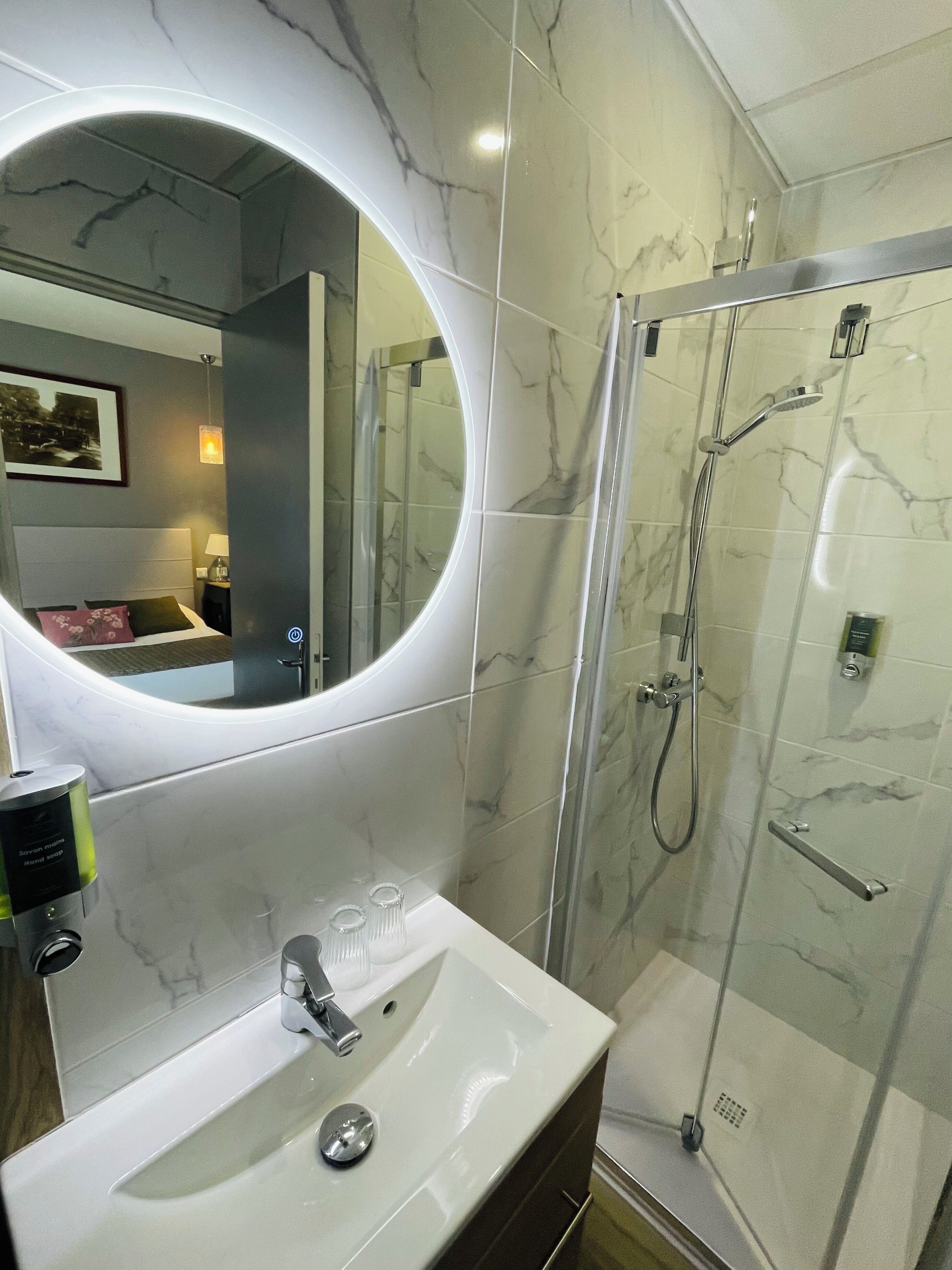 Une salle de bain avec un mur recouvert de carreaux de marbre, un miroir rond, un lavabo blanc et une douche à parois de verre.