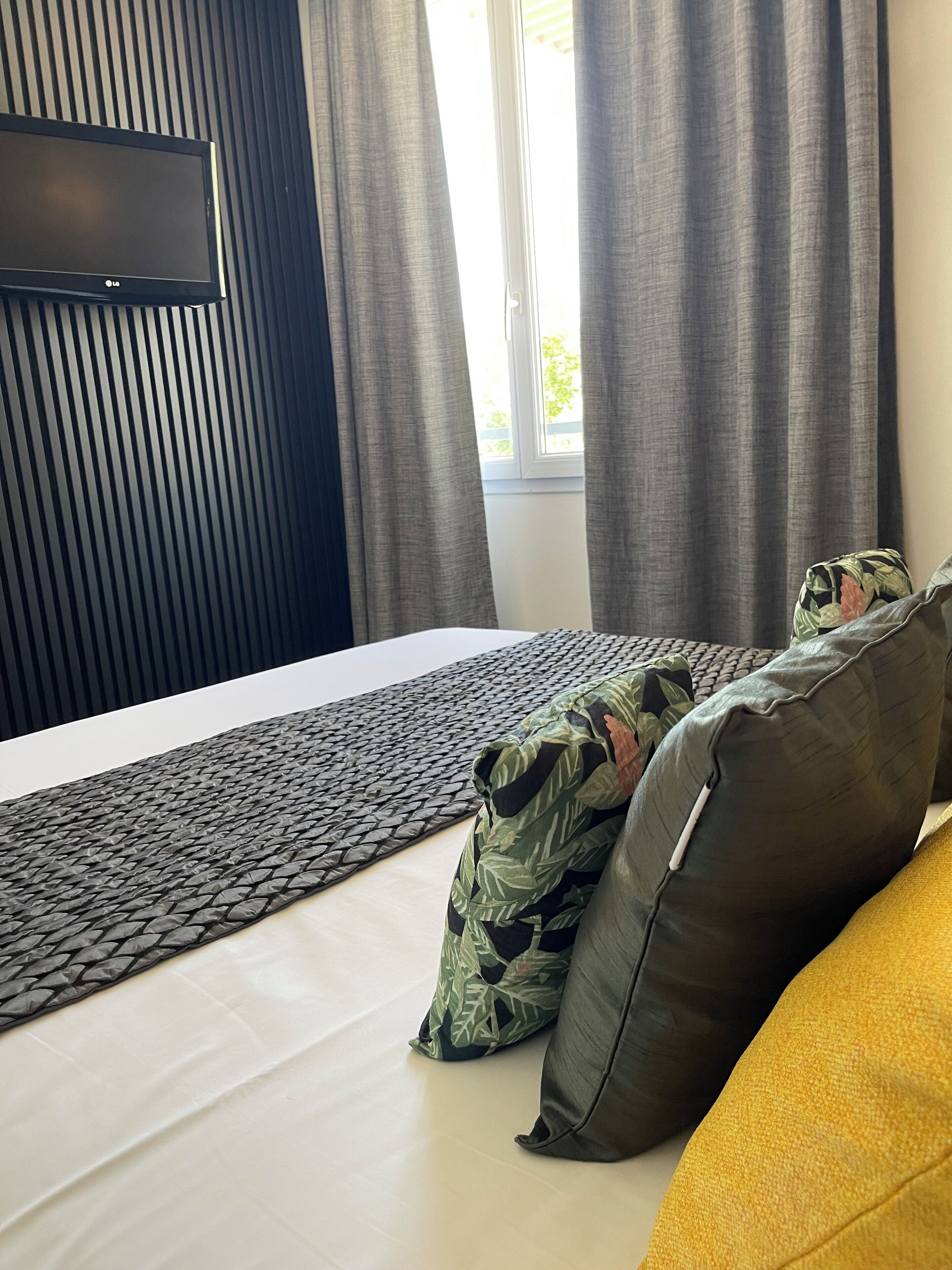 A modern hotel room with a large bed, a flat-screen TV mounted on the wall, and a view of the outdoors through the window.