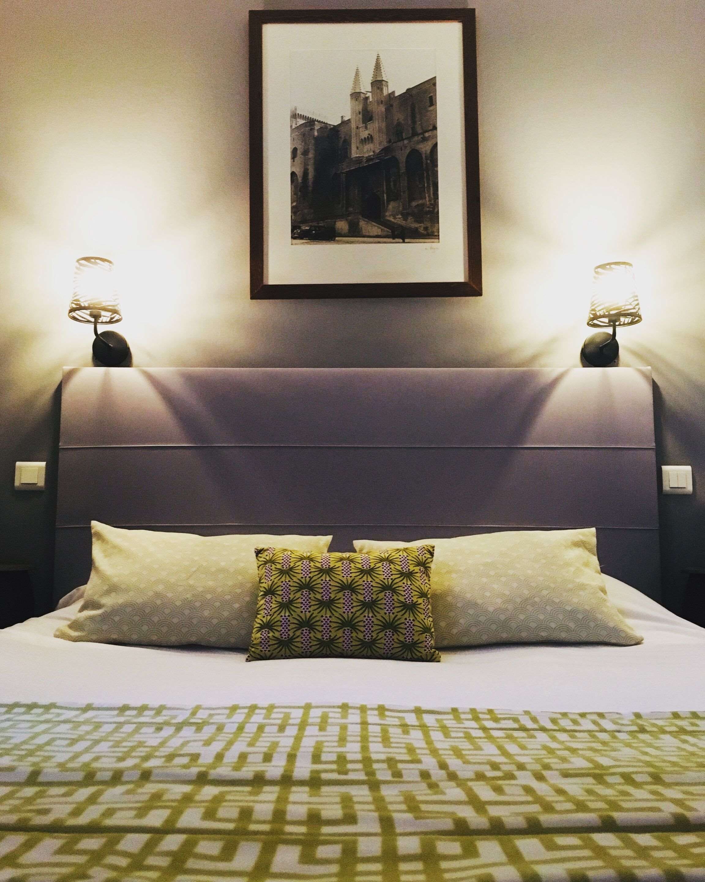 Una cama bien tendida con almohadas estampadas, un cuadro enmarcado encima y dos lámparas de pared a cada lado.