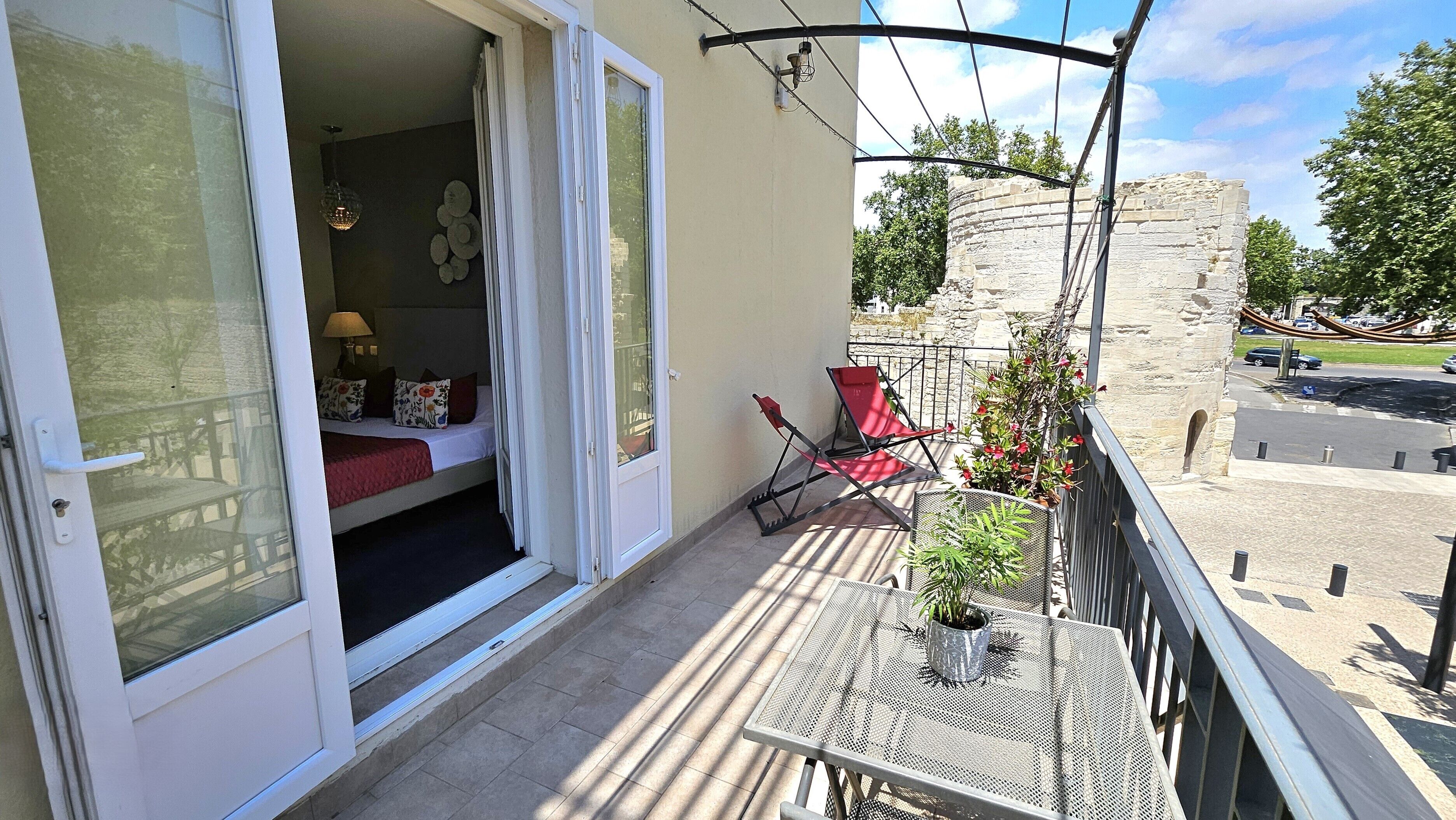 Un balcon aménagé avec un lit, une table et des chaises, donnant sur un mur de pierre et une rue.