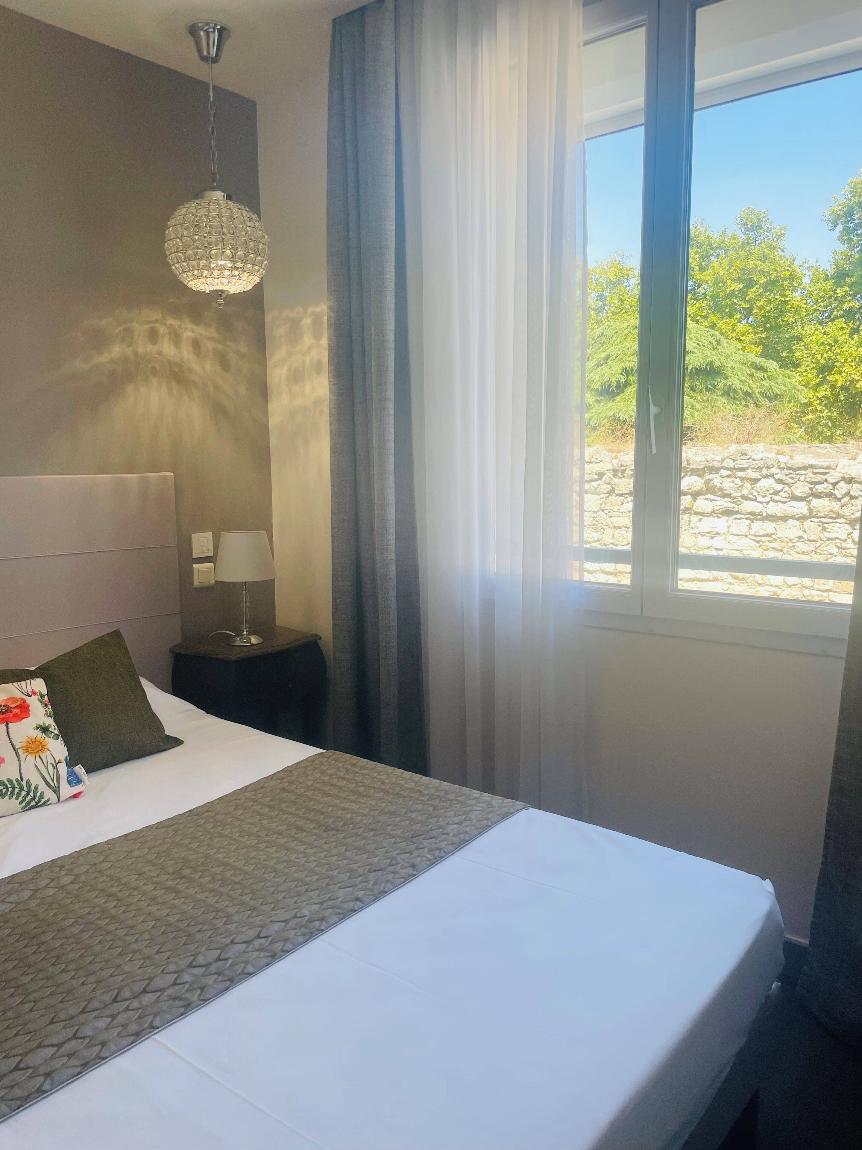 Une chambre d’hôtel avec un lit, une lampe de chevet, une suspension décorative et une fenêtre donnant sur un paysage verdoyant.
