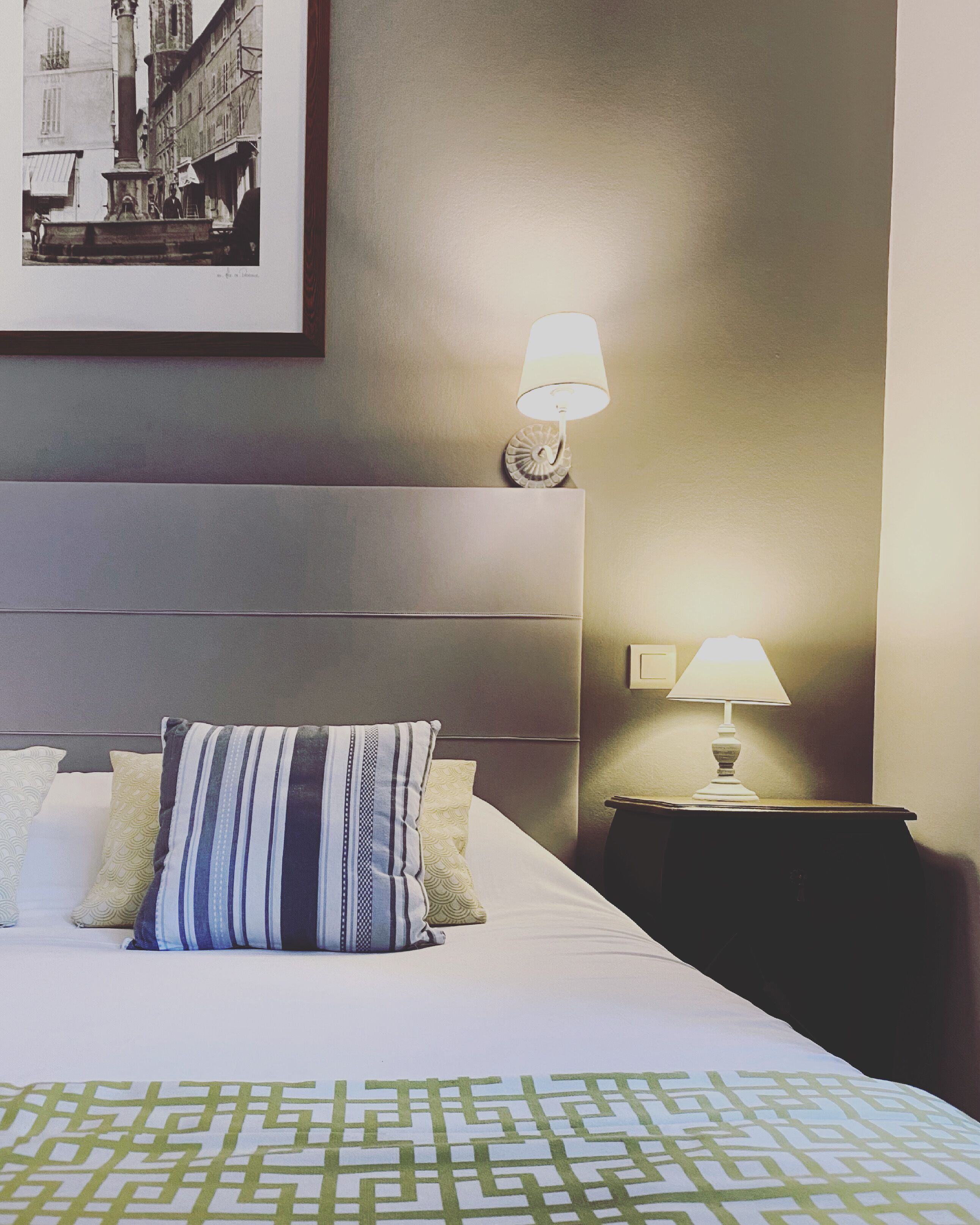 A hotel room with a bed, pillows, a nightstand, two wall-mounted lamps, and a framed picture on the wall.