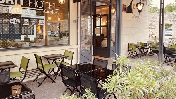 A BRIT HOTEL Comfort exterior with outdoor seating, a glass door entrance, and a stone wall backdrop. - Brit Hotel Acacias (Arles)