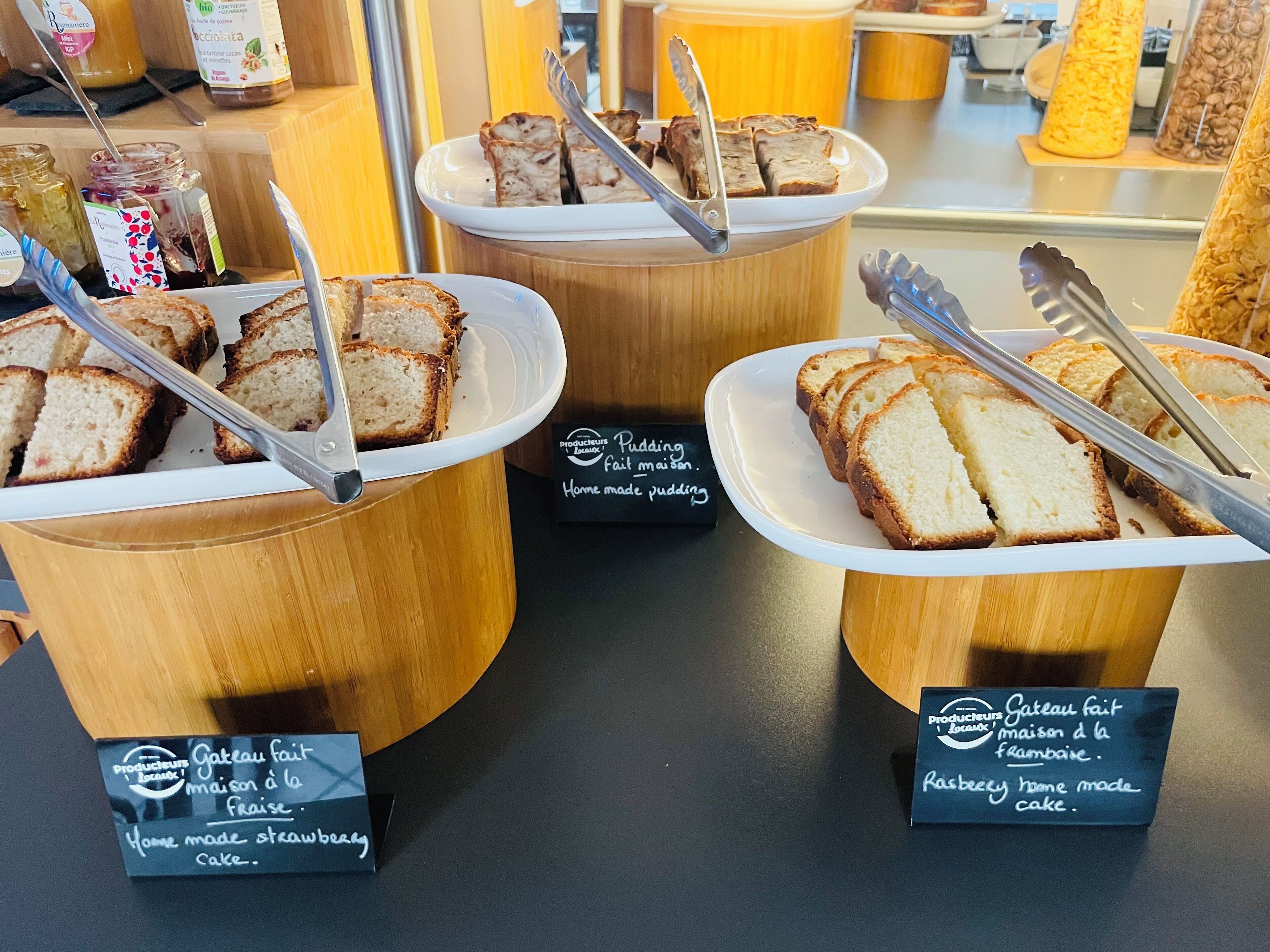Trois récipients en bois contenant des parts de gâteau et des pinces, chacun étant étiqueté avec le nom d’un type de gâteau différent.