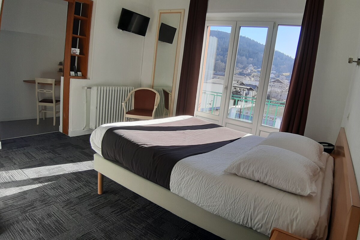 A hotel room with a bed, a chair, a desk, a TV, and a window with a view of a town and mountains.