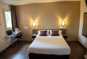 A hotel room with a large bed, two bedside tables, a desk with a laptop, and two wall-mounted lights.