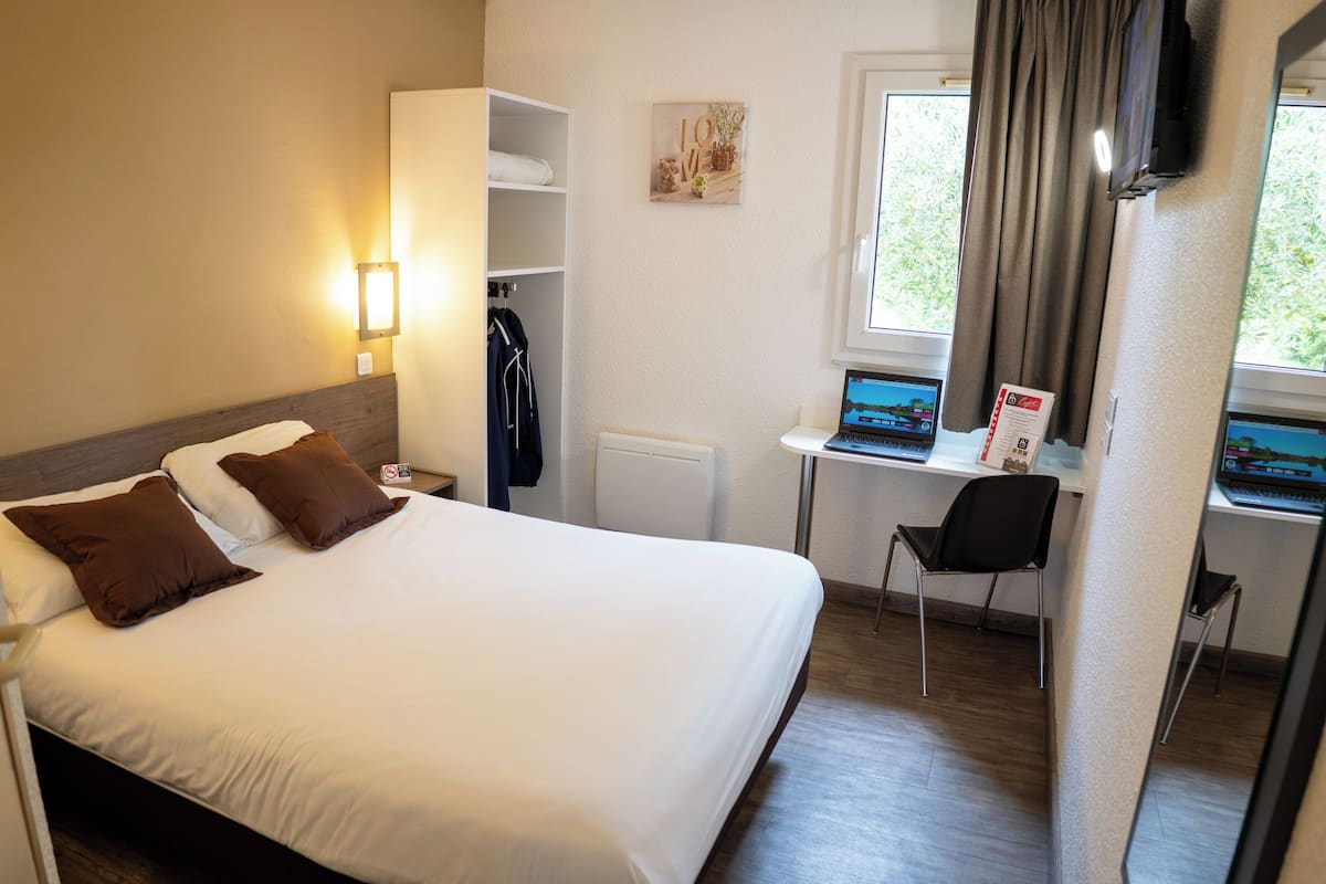A hotel room with a bed, a desk with a laptop, a chair, and a coat rack.