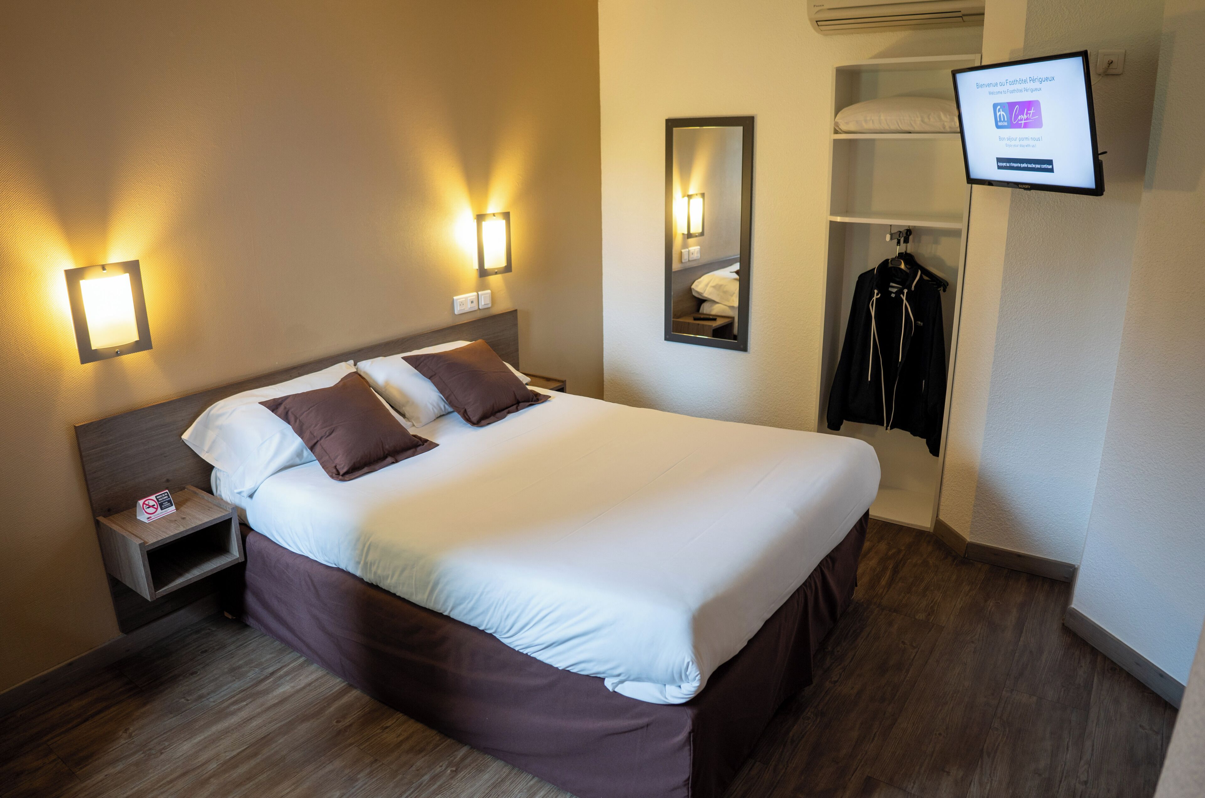 Una habitación de hotel con cama, mesita de noche, televisor montado en la pared y perchero.