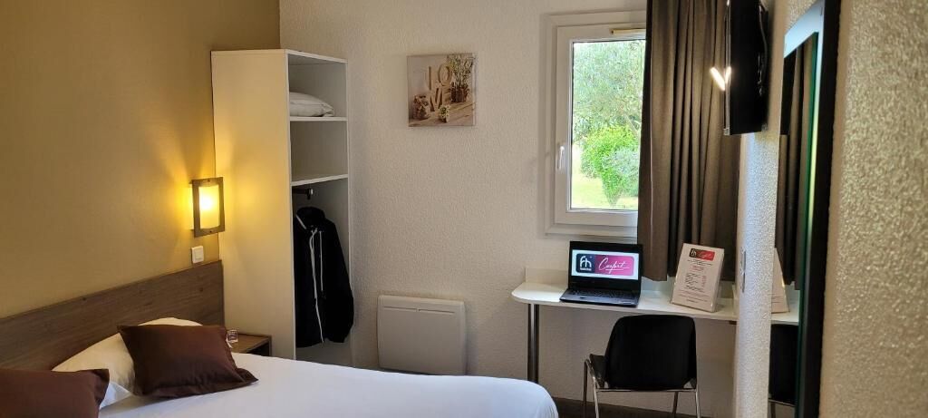 A hotel room with a bed, a desk with a laptop, a chair, a coat rack, and a window with curtains.