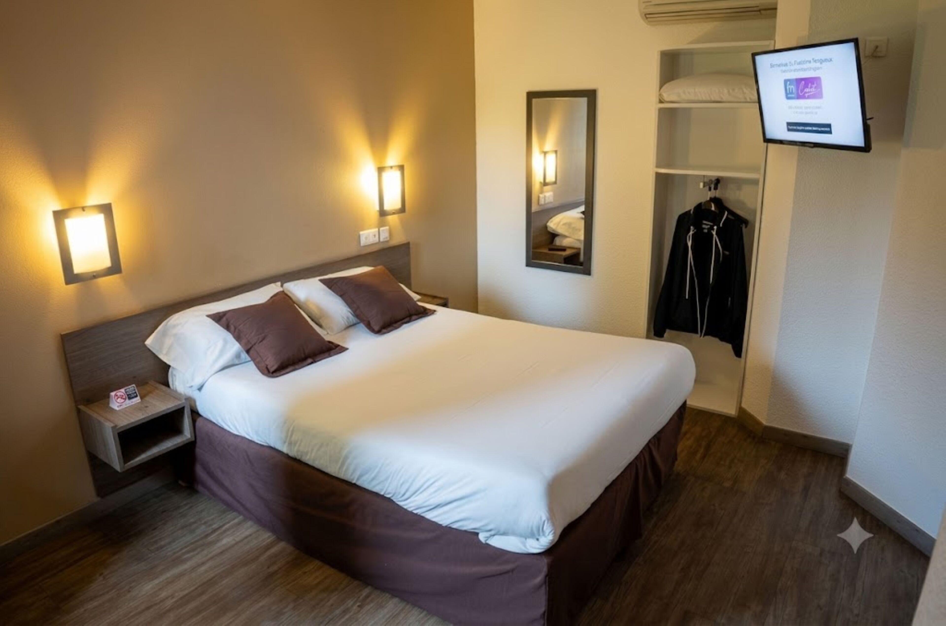 A hotel room with a bed, bedside tables, a mirror, a coat rack, and a TV screen displaying a message.