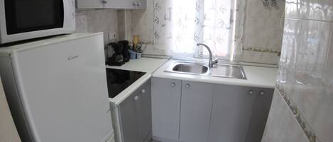 A compact kitchen with a microwave, refrigerator, sink, and a window with decorative curtains.