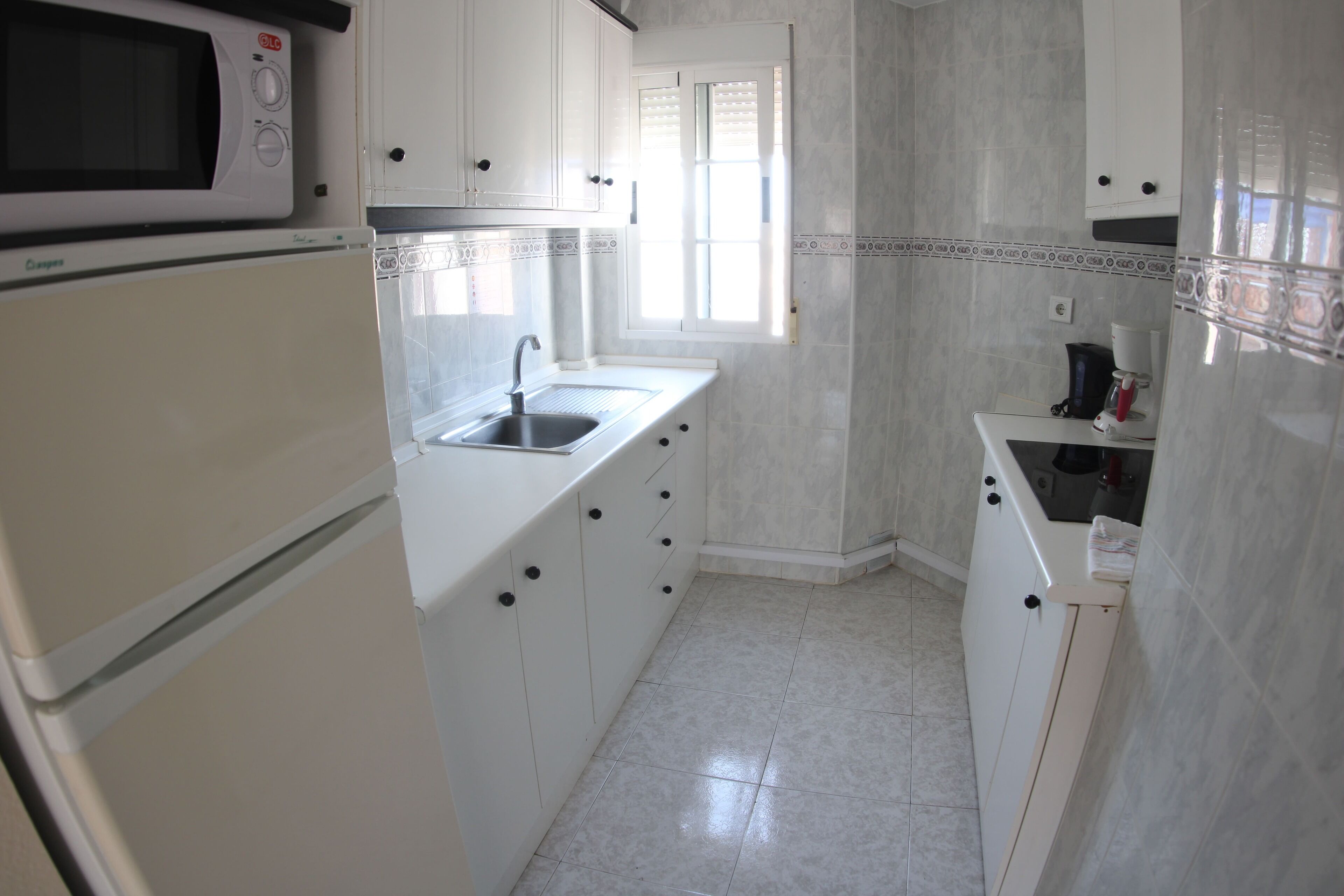 Una cocina con gabinetes blancos, microondas, fregadero y ventana.