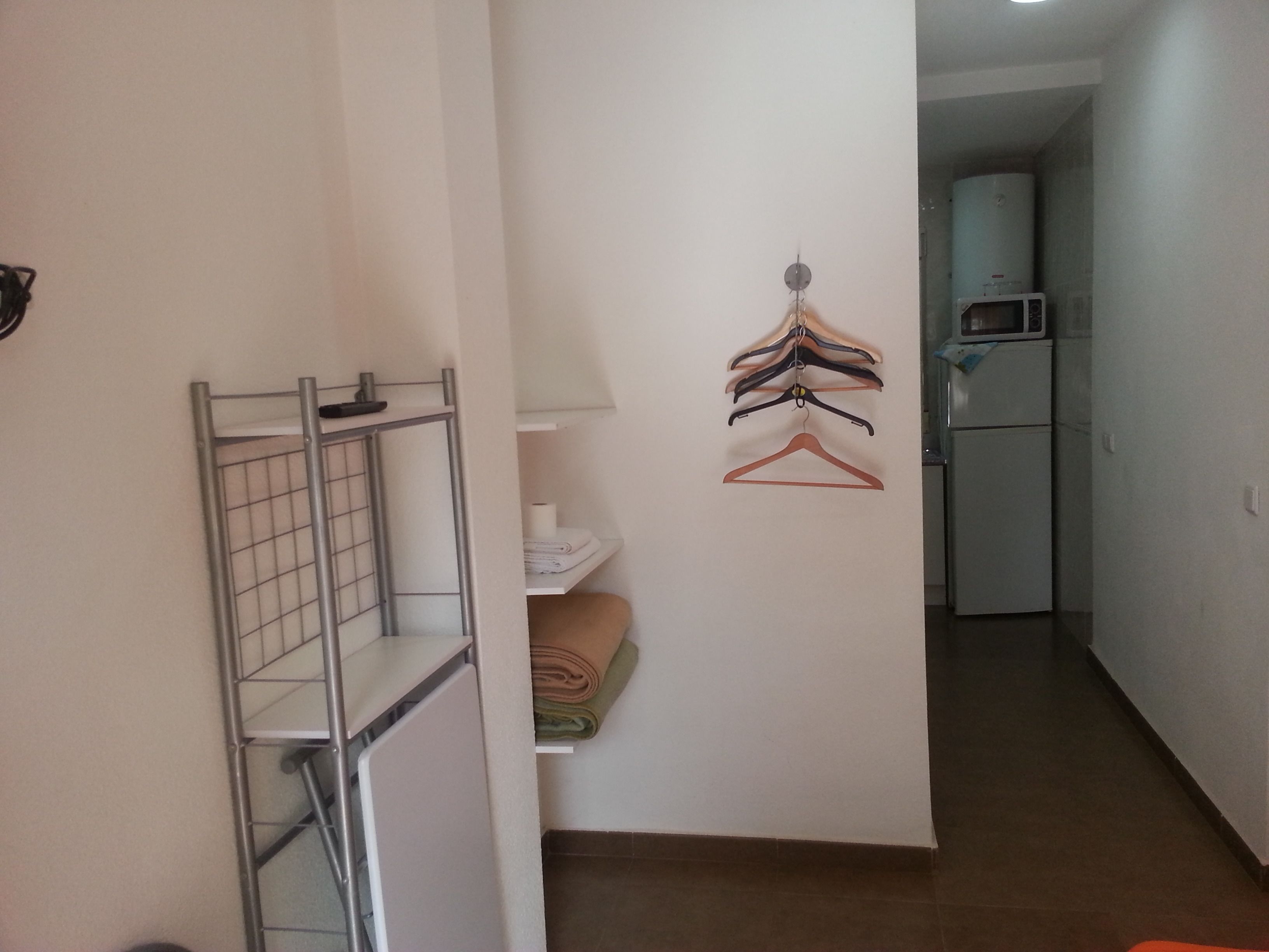 A narrow hallway with a metal drying rack, a wall-mounted clothes rack with hangers, a folded towel, and a white refrigerator.
